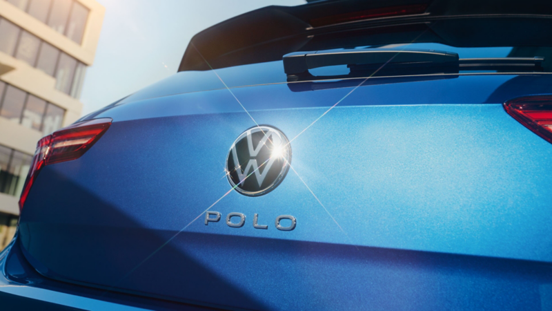Die geschlossene Kofferraumklappe eines blauen VW Polo mit VW-Logo, in dem sich die Sonne spiegelt.