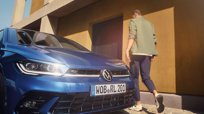 Mężczyzna podchodzi do drzwi kierowcy niebieskiego VW Polo, które stoi przed żółtym budynkiem