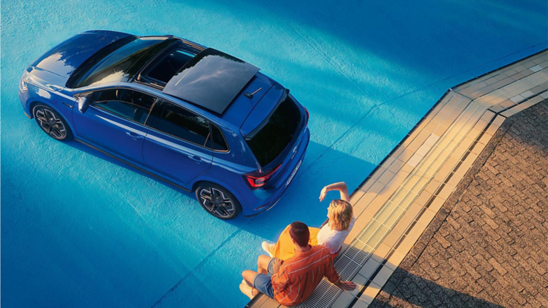 Ein blauer VW Polo steht in einem leeren Pool, ein Pärchen sitzt am Rand. Blick schräg von oben auf das optionale Panorama Ausstell-/Schiebedach.