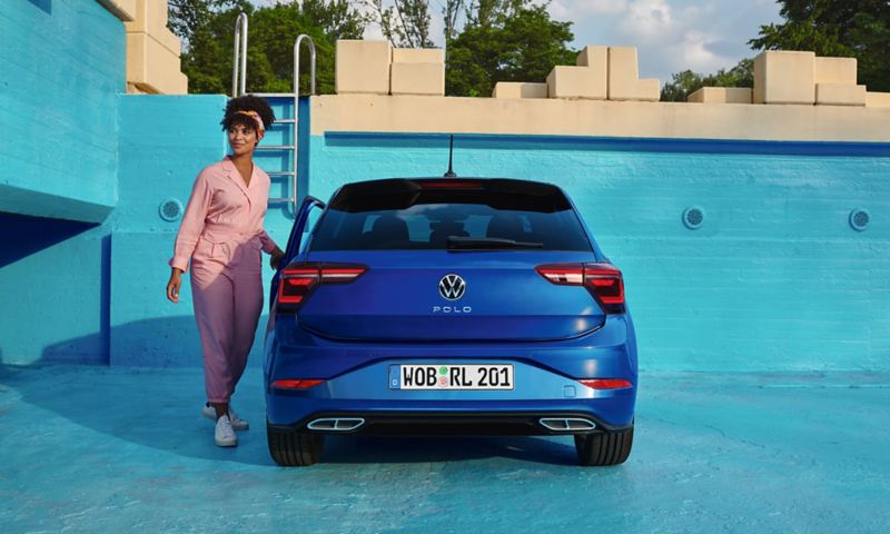 Blick auf das Heck eines blauen VW Polo, gepark tin einem leeren Pool. Eine Frau im rosa Overall öffnet die Fahrertür. 