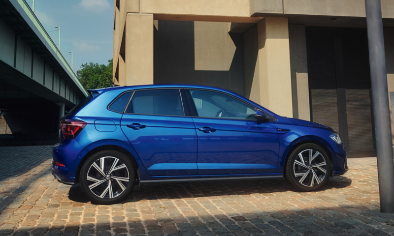 View of the passenger side of a blue VW Polo, parked next to a high-rise building.