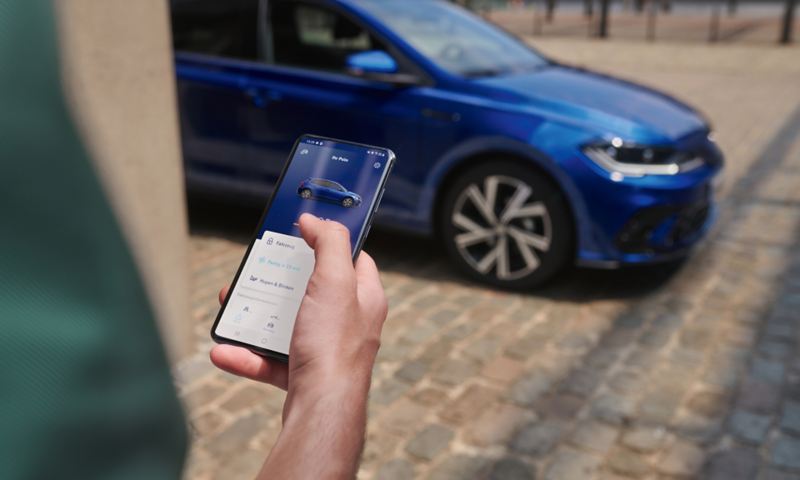Blik på en mobiltelefons display med bildata fra Polo, i baggrunden holder en blå VW Polo parkeret.