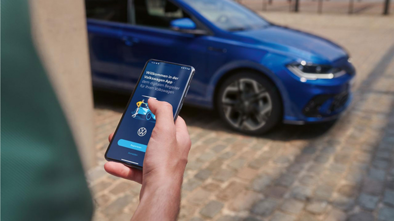 Ein blauer VW Polo steht auf einem Parkplatz. Eine Person prüft dessen Fahrdaten auf dem Smartphone.
