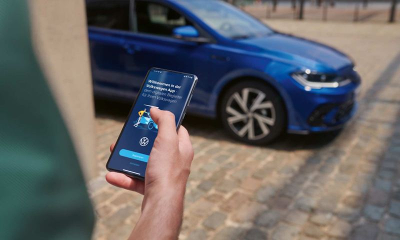 Vue sur l’écran d’un téléphone portable avec les données de la Polo, en arrière-plan la VW Polo bleue, garée.