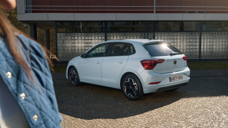 Une Polo Edition 50 bleu clair est garée en travers devant un bâtiment et une femme se tient à côté.
