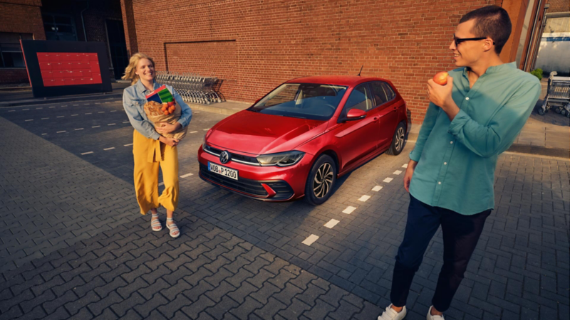 Ein roter VW Polo steht auf einem Parkplatz vor einem Supermarkt, ein lachendes Paar steht mit Einkäufen daneben.