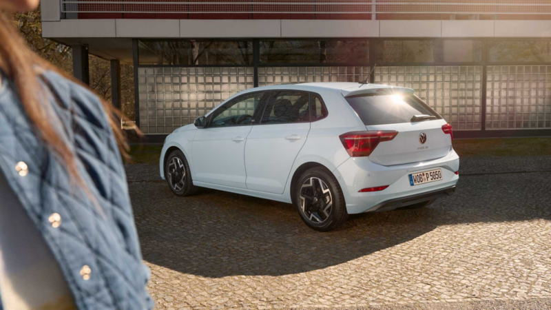 Ein hellblauer Polo EDITION50 parkt seitlich vor einem Gebäude und eine Frau steht vor ihm.