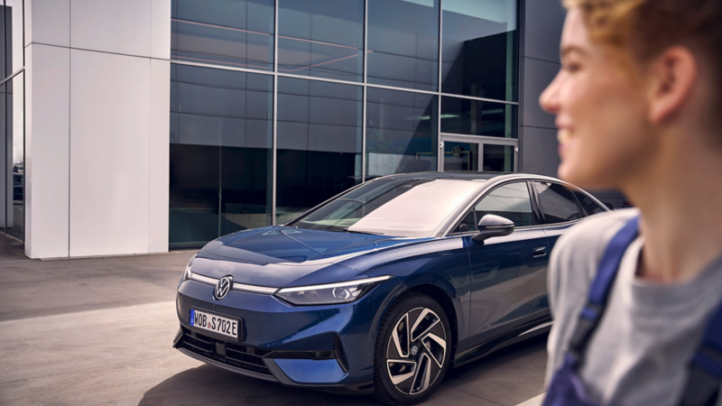 Eine Servicemitarbeiterin steht vor einem blauen VW ID.7, der vor einem VW Händlergebäude parkt.
