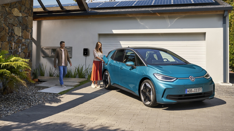 Eine Frau lädt einen Volkswagen ID.4 vor einem modernen Haus mit Solarpanels auf dem Dach; ein Mann kommt dazu.