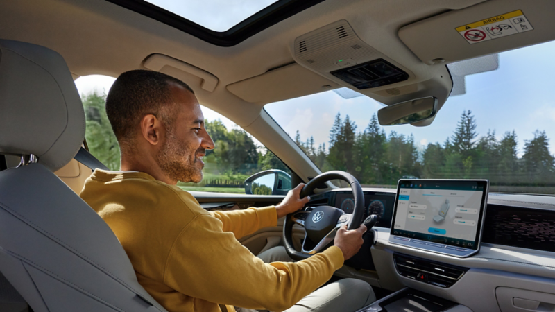  Un homme souriant et barbu est assis au volant d'une Volkswagen Passat. Il porte un pull jaune et regarde de côté. On peut voir le volant et le tableau de bord numérique, ainsi qu'un écran tactile central qui affiche un schéma des sièges avec des commandes de réglage.
