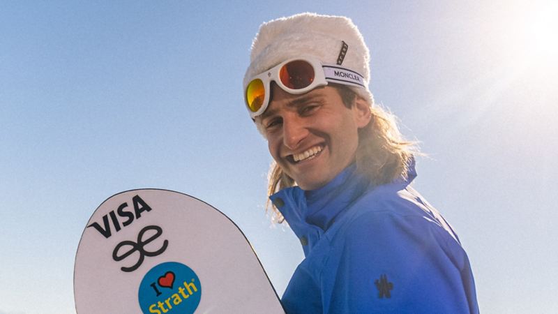 Pat Burgener steht mit seinem Snowboard in der Hand und lächelt in die Kamera