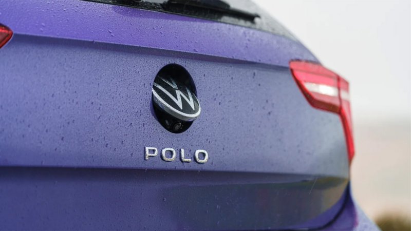 A man walks to the driver’s door of a blue VW Polo standing in front of a yellow building.