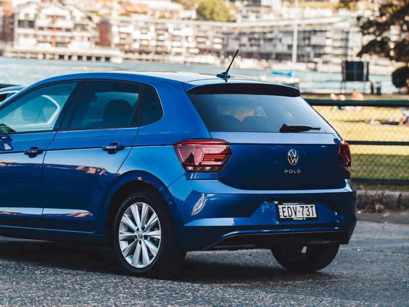 A blue Volkswagen Polo car is parked by a city park near a river