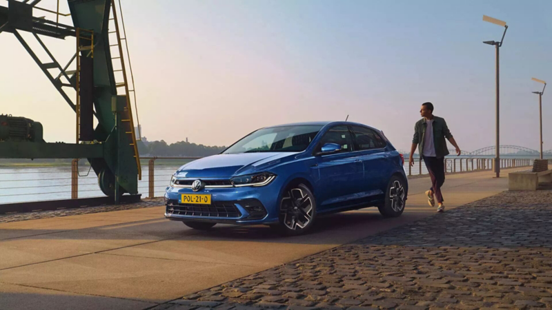 Schuin vooraanzicht van blauwe Volkswagen Polo op industrieterrein, een man loopt achter de auto