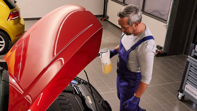 Técnico de Volkswagen revisa motor de auto rojo, sosteniendo líquido de mantenimiento en taller oficial.