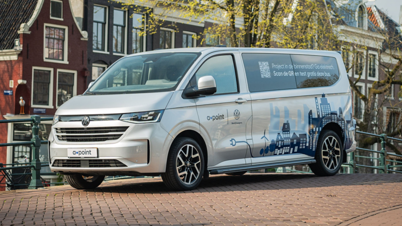 Grijze Volkswagen e-Transporter van a·point op een brug. Tekst: Project in de binnenstad? Ga elektrisch. Scan de QR en test gratis deze bus.