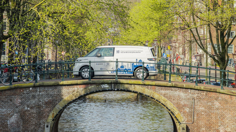 Elektrische ID. Buzz Cargo van a·point op een Amsterdamse boogbrug. Tekst: Project in de binnenstad? Ga elektrisch. Scan de QR en test gratis deze bus.