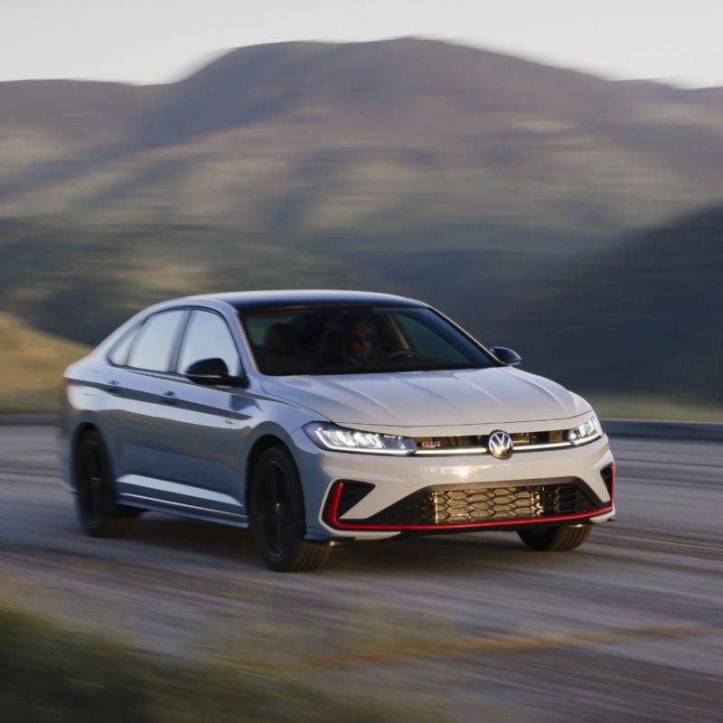 Une Volkswagen Jetta GLI 2025 argentée en mouvement sur une route, avec des collines montagneuses floues en arrière-plan.