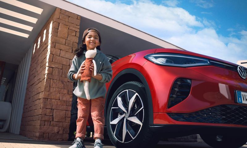 Inquadratura ravvicinata dal basso di una bambina sorridente mentre tiene un mano un pupazzo e dietro di lei parcheggiata in un garage una vettura Volkswagen.