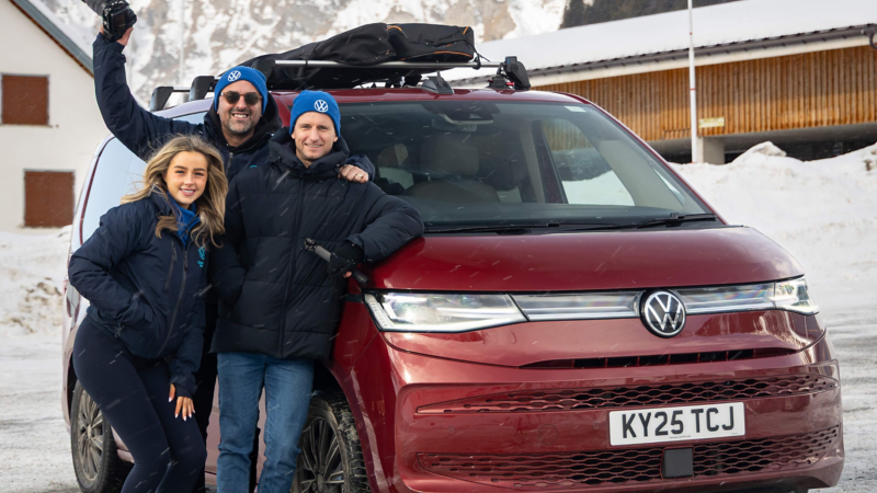 Three people stand cheerfully in front of a maroon Volkswagen Multivan, surrounded by snow. They're dressed in winter clothing and hats, conveying a joyful tone.
