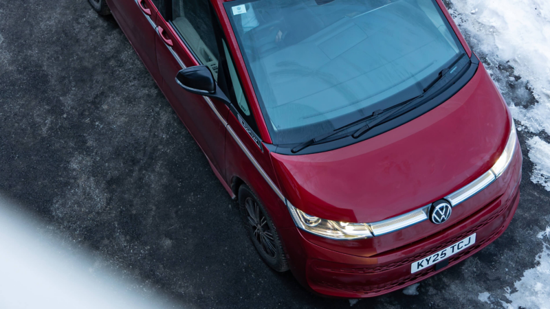 A red Volkswagen Multivan is parked on a snowy, icy road. The car's sleek design contrasts with the white snow, creating a vibrant and clean visual.