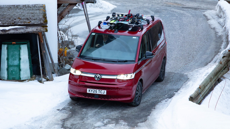 Red Volkswagen Multivan drives on a snowy road by a cabin. Roof holds skis and snowboards. Scene conveys winter adventure and travel in icy conditions.