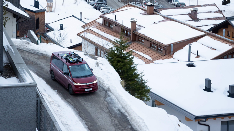 Red Multivan with skis on top driving on snowy road in a mountain village, surrounded by snow-covered rooftops and evergreen trees. Winter scene.
