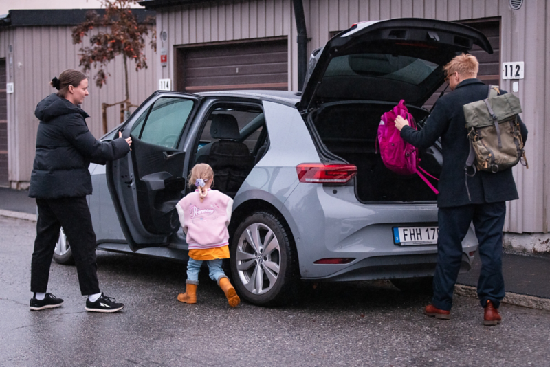 En grå Volkswagen ID.3 står parkerad vid en gata, en familj är påväg in och packar bagageutrymmet