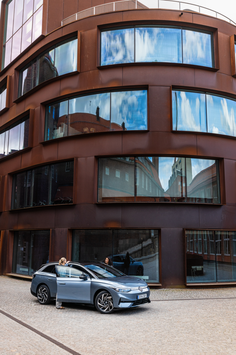 Två personer kliver ur en ID.7 Tourer som står parkerad framför en modern stadsbyggnad med rundad fasad och stora fönster. 