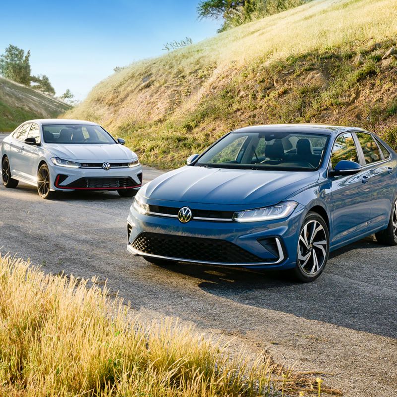 Vue de face de la Volkswagen Jetta 2025 bleue et Jetta GLI blanche mettant en valeur son nouveau design extérieur avec des collines pittoresques en arrière-plan.