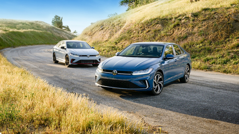 Front view of the blue 2025 Volkswagen Jetta and white Jetta GLi showcasing its new exterior design with scenic hills in the background.