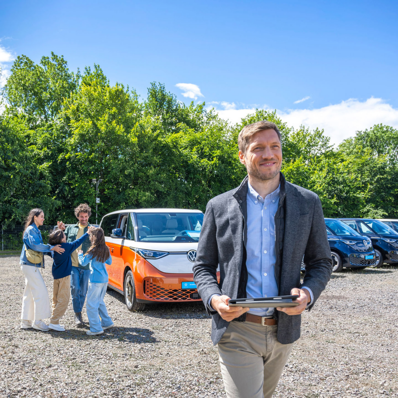 ProfiPartner auf einem Autohof, ein Mann mit Tablet in der Hand im Vordergrund, im Hintergrund parken verschiedene Volkswagen Nutzfahrzeuge Modelle.