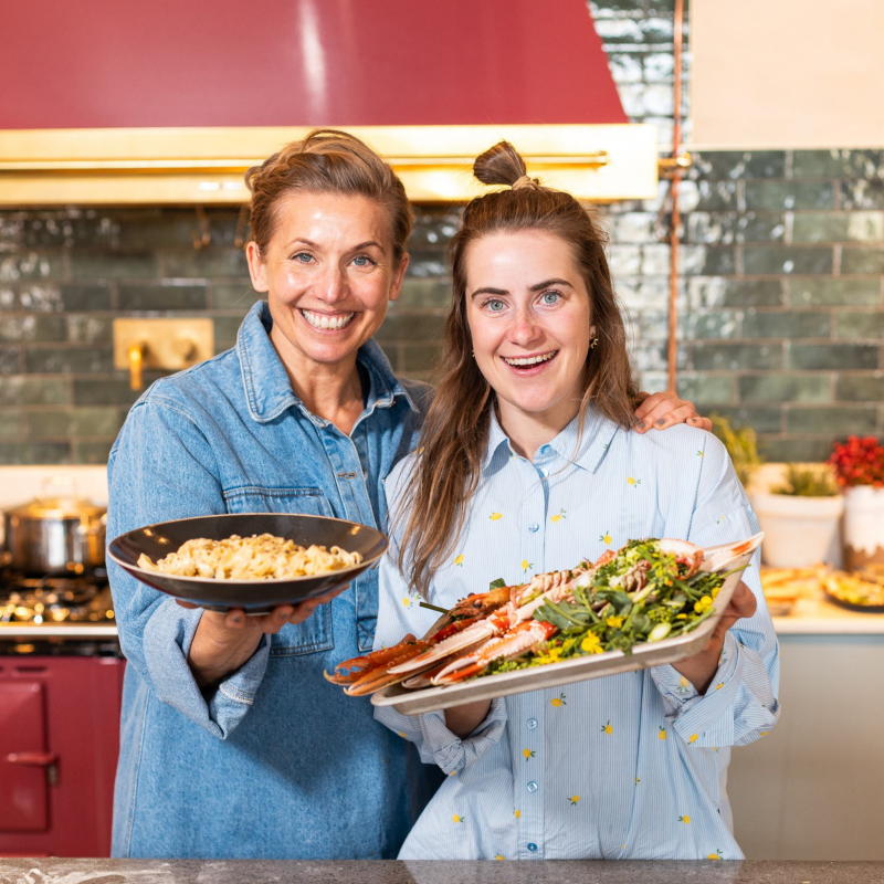 Ebba andersson och Tina Nordström står i ett kök och visar upp maten de lagat