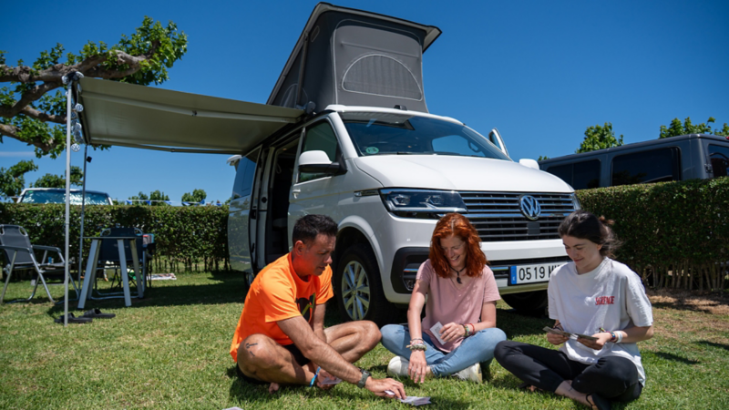 Una familia jugando a cartas sentada en el césped, delante de un VW California.