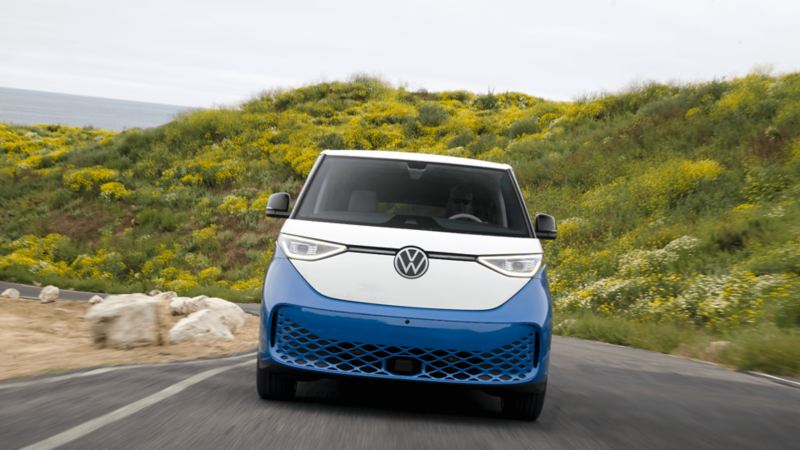 Un Volkswagen ID. Buzz 2025 bleu et blanc roule sur une route avec des collines verdoyantes en arrière-plan.