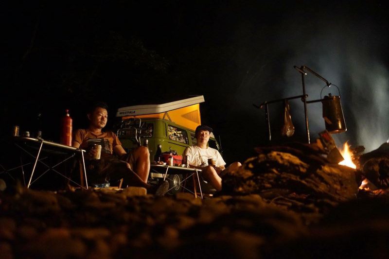 車主小雷在夜晚營火前與友人用餐笑談的照片，遠方停著掀起黃色車頂帳的綠色T2 Westfalia