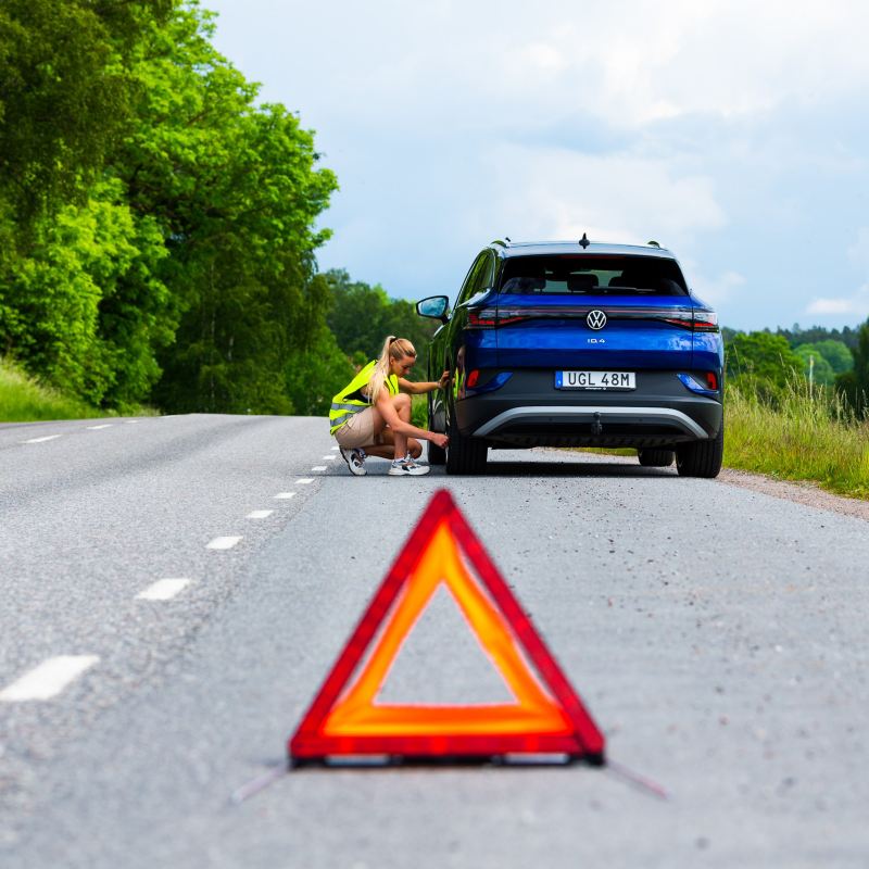 Mobilitetsgaranti - En varningstriangel på vägen, kvinna med reflexväst sitter vid en bil
