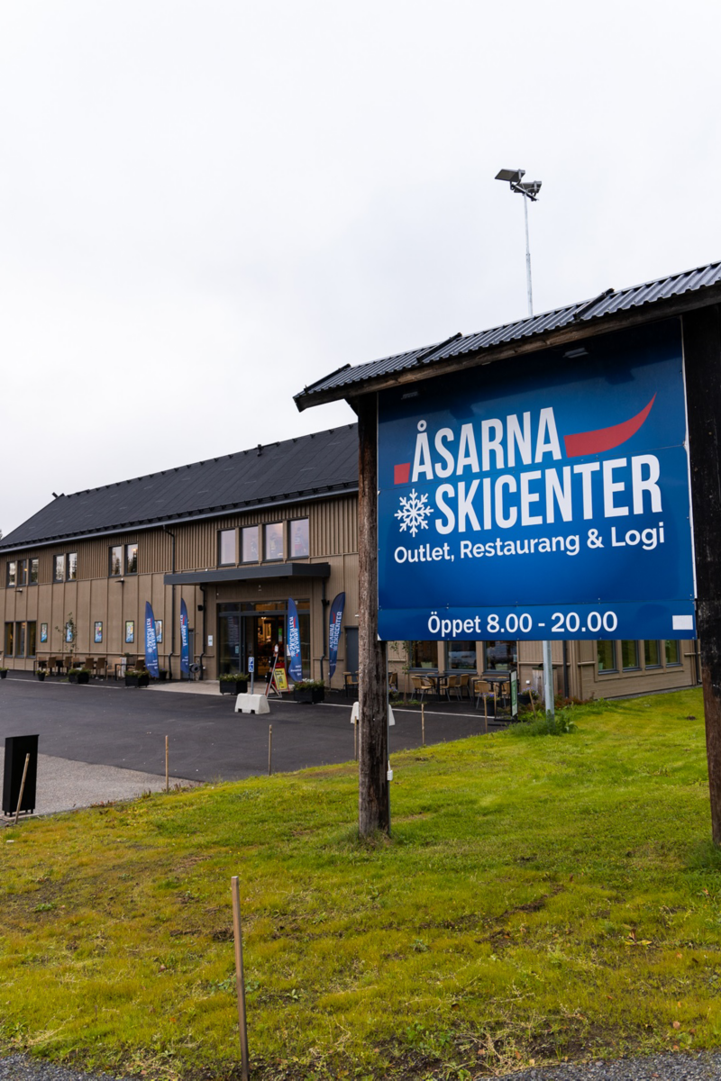 Skylt i gräset med texten "Åsarna Skicenter", i bakgrunden står en byggnad