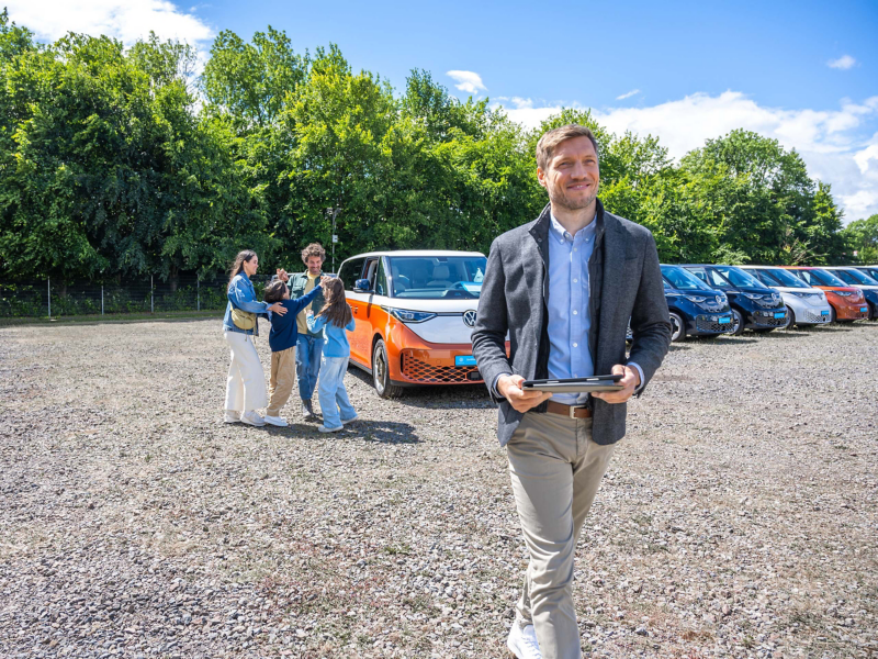 ProfiPartner auf einem Autohof, ein Mann mit Tablet in der Hand im Vordergrund, im Hintergrund eine sich freuende Familie und verschiedene parkende Volkswagen Nutzfahrzeug Modelle.