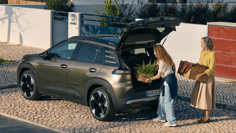A woman loading a box with fern leave plants into the boot of a VW ID. Cross. A woman carrying a box with clothes inside is standing behind.