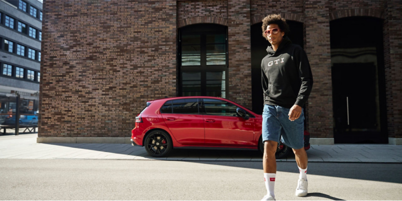 Man in GTI sweatshirt walking past VW GTI on city street.