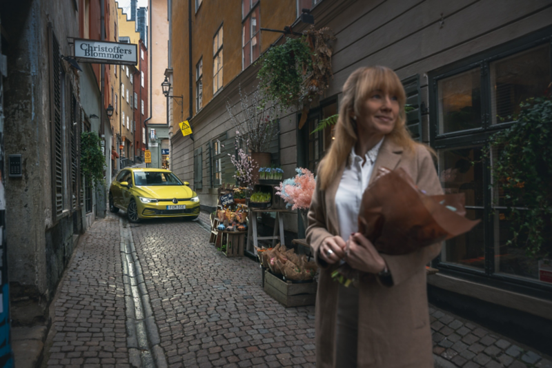 En person står i en gränd vid en blomsterbutik. I bakgrunden står en gul Volkswagen.