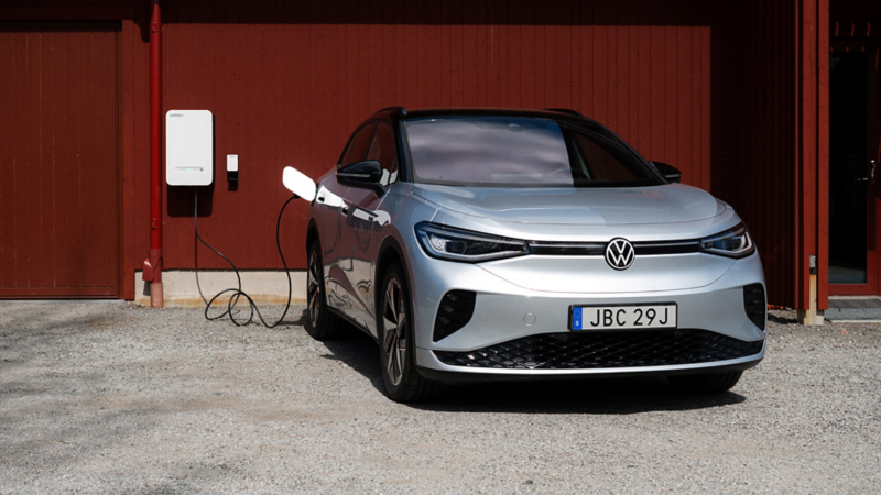 En VW ID.-bil står parkerad vid ett rött hus. En laddningskabel är inkopplad.
