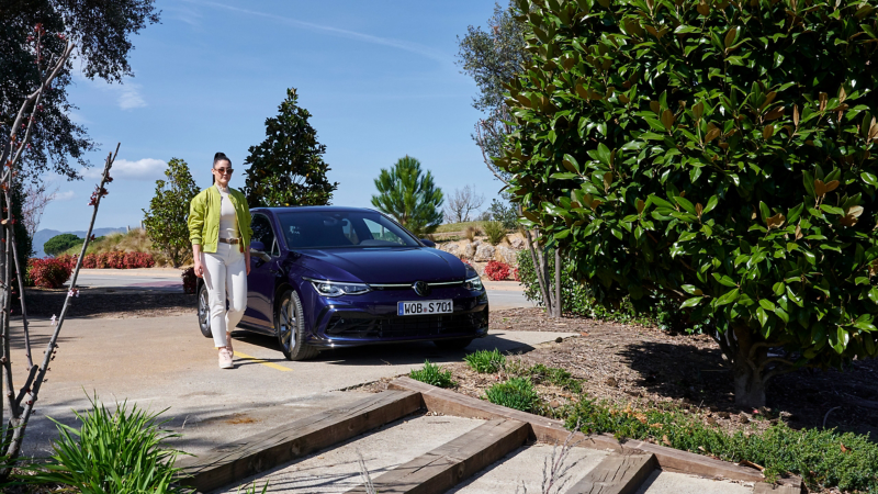 Woman walks next to parked VW Golf, Mediterranean landscape in background