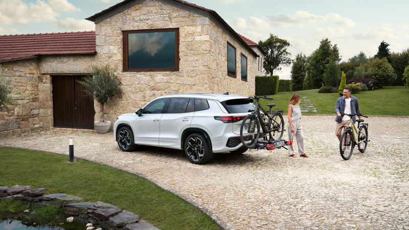 Una mujer de pie junto a un Volkswagen Tayron con una bicicleta en el portabicicletas, llega un hombre empujando una bicicleta