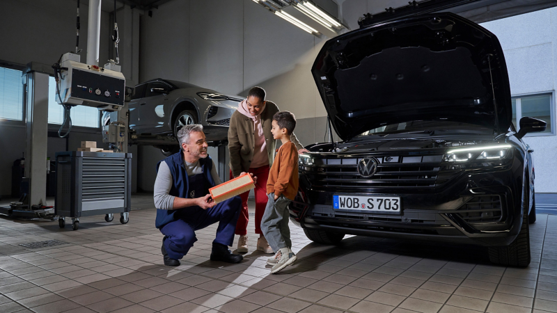 Un empleado del servicio técnico de VW muestra a los clientes un filtro de aire - VW Touareg con el capó abierto al fondo
