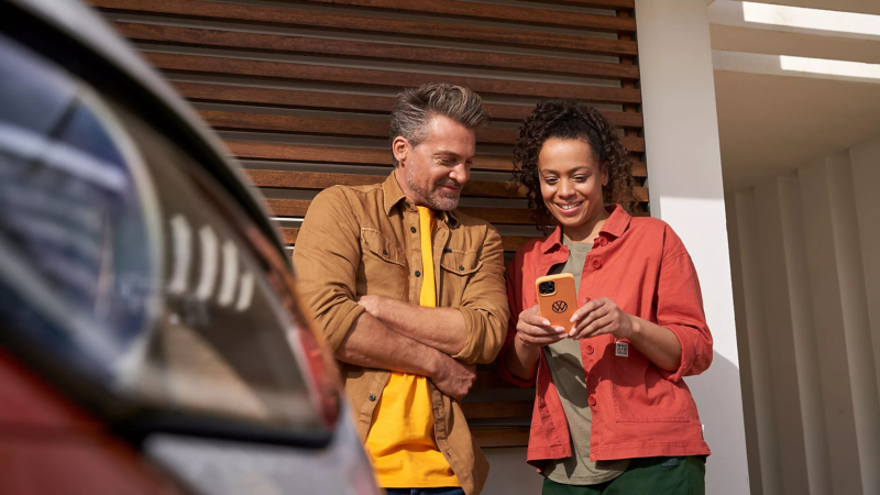 Vrouw en man kijken naar de telefoon die de vrouw in de handen heeft, ze leunen tegen een garage, op de voorgrond een gedeelte van een auto