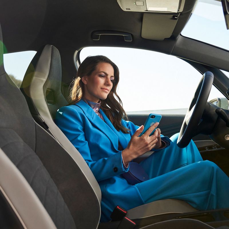 Eine Frau sitzt in einem VW ID.3 und schaut auf ihr Handy – Hilfe zu Apps und digitalen Diensten