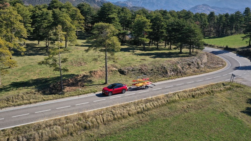 Une voiture Volkswagen rouge tire une petite remorque avec deux kayaks sur une route de campagne sinueuse, entourée d'arbres.