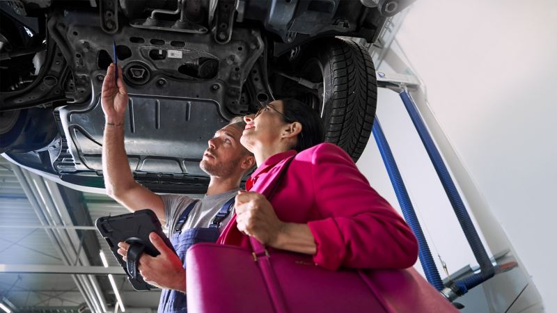 Un collaborateur d’atelier Volkswagen et une cliente regardent le dessous d’une voiture Volkswagen